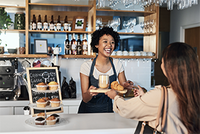 Barista hinter dem Tresen eines Cafés reicht einer Kundin ein Tablett mit Kaffee und Croissant.