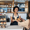 Barista hinter dem Tresen eines Cafés reicht einer Kundin ein Tablett mit Kaffee und Croissant.