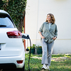 Eine Frau steht im Garten neben einem weißen Elektroauto, das gerade an einer Ladestation angeschlossen ist.