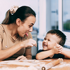 Eine junge Au-pair-Betreuerin sitzt gemeinsam mit einem lachenden Kind am Tisch. Beide lächeln sich an und verbringen eine fröhliche Zeit miteinander. Das Bild illustriert das Thema Au-pair-Aufenthalt und dessen steuerliche Grundlagen.