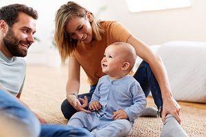 Ein Mann und eine Frau sitzen auf dem Boden. Sie schauen ein Baby an, das zwischen ihnen auf dem Boden sitzt. Alle lächeln und wirken glücklich. Die Frau streck ihren Zeigefinger an die Hand des Babys.