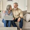 Ein Mann und eine Frau, beide grauhaarig, sitzen nebeneinander auf einem Sofa und lachen. Auf dem Schoß der Frau liegt ein Laptop.