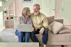 Ein Mann und eine Frau, beide grauhaarig, sitzen nebeneinander auf einem Sofa und lachen. Auf dem Schoß der Frau liegt ein Laptop.