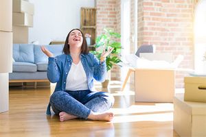 Eine Frau in Jeans und Hemd sitzt im Schneidersitz auf dem Boden ihre Wohnung. Sie lacht, schaut gen Decke und hält ihre Hände freudig nach oben. Um sie herum befinden sich Umzugskartons, teilweise schon ausgepackt. Im Hintergrund steht eine Couch.