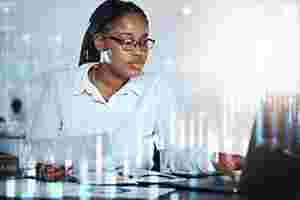 Eine Frau mit Brille sitzt am Schreibtisch und schaut auf ihren Laptop. Über das gesamte Bild laufen in hellem weiß Zahlen und Diagramme. 