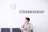 Fehlerhafte Übermittlung der vermögenswirksamen Leistungen Ein Mann schaut ungeduldig auf eine Uhr an der Wand. An der Wand steht „Steuerbescheid?“