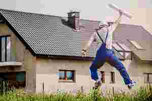 Das Bild zeigt die Rückensicht eines Mannes in Trägeranzug und Schutzhelm. Er befindet sich vor einem großen Haus. Er springt in die Luft und hält in der einen Hand eine Rolle Papier, die er nach oben streckt.