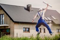 Neue Sonderabschreibung für Mietwohnungen Das Bild zeigt die Rückensicht eines Mannes in Trägeranzug und Schutzhelm. Er befindet sich vor einem großen Haus. Er springt in die Luft und hält in der einen Hand eine Rolle Papier, die er nach oben streckt.