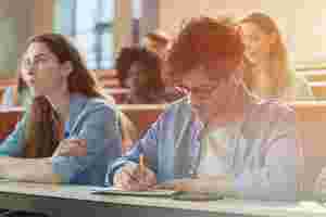 In einem Vorlesungsaal sitzen Studenten, dabei sind zwei deutlich zu erkennen. Eine Studentin schaut nach vorne, ein anderer Student schreibt etwas auf. Der Hintergrund ist verschwommen.