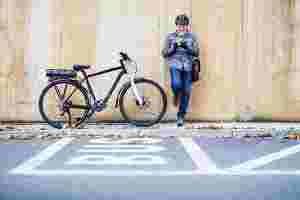 Ein älterer Mann steht auf einem Bürgersteig und lehnt sich mit dem Rücken an eine Wand, ein Bein ist angewinkelt. Er tippt etwas auf seinem Smartphone. Er trägt einen Fahrradhelm und eine Umhängetasche. Links neben ihm steht ein Elektro-Fahrrad. Auf der Straße vor ihm ist eine Spur mit der Aufschrift „BUS“ eingezeichnet.