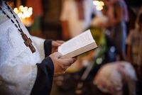 Die Kirchensteuer als Sonderausgaben absetzen Ein Priester im hellen Gewand ist von der Seite zu sehen. Er hält die Bibel aufgeschlagen in seiner Hand. Im Hintergrund findet ein Gottesdienst statt.