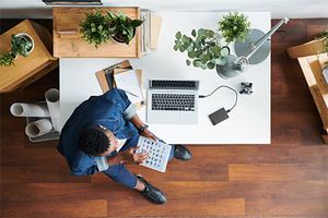 Das Bild zeigt aus der Vogelperspektive einen Mann in Jeans und Hmed halb auf einem Schreibtisch sitzen. Er hält ein Tablet in der Hand. Auf dem Schreibtisch befinden sich ein Laptop, ein Handy, Stifte, Papierunterlagen und Pflanzen. Neben dem Schreibtisch stehen ein Korb mit großen Papierrollen und weitere Pflanzen.