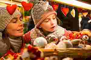 Eine Frau mit Wollmütze und Schal hat ihre kleine Tochter, die ebenfalls eine Wollmütze trägt, auf dem Arm. Beide sind auf dem Weihnachtsmarkt und schauen sich an einem Stand Weihnachtsschmuck an. Im Vordergrund hängen rote Herzen an einer Girlande. Das Kind staunt über die schönen Sachen und greift nach einer Christbaumkugel.
