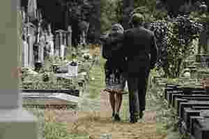 Eine Frau und ein Mann in schwarzer Kleidung laufen tagsüber über einen Friedhof zwischen Gräbern entlang. Sie sind von hinten zu sehen; der Mann hat seinen Arm um die Frau gelegt. Rechts und links von dem Paar befinden sich Gräber, Blumen und Gestecke.