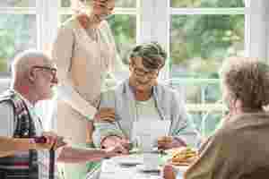 Eine Gruppe von drei Senioren sitzt an einem Frühstückstisch. Eine Seniorin ist von vorne zu sehen, sie hält ein Blatt Papier in der Hand und blickt lächelnd darauf. Eine junge Pflegerin steht neben ihr und legt ihr de Hand auf die Schulter.