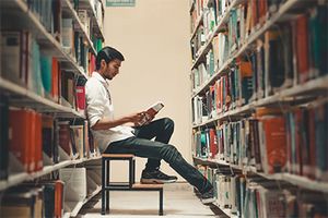 Ein junger Student ist in der Universitätsbibliothek und sitzt zwischen zwei großen Bücherreihen auf einem kleinen Tritt. Er trägt Jeans und Hemd und hält ein aufgeschlagenes Buch in der Hand, das er liest. Der Student ist von der Seite abgebildet, die Bücherreihen sind im Vordergrund verschwommen.