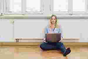 Eine junge Frau sitzt auf dem Boden vor der Heizung und schaut auf ihren Laptop.