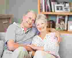 Ein älteres Ehepaar sitzt gemeinsam auf der Couch und lächelt in die Kamera.