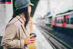 Eine Frau mit langen Haaren, Mütze und Mantel hält einen Coffee-to-go-Becher in der Hand und steht am Bahnhof. Sie schaut auf die Gleise und wartet auf ihren Zug.