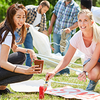Eine Gruppe junger Menschen bereitet im Freien Banner vor. Zwei Frauen im Vordergrund knien auf dem Gras, eine hält einen Becher, die andere malt mit einem Pinsel auf das Banner.