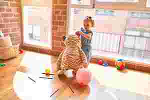 Ein kleines Kind steht im Kindergarten vor einem großen Fenster. Sie hält einen großen Stoffbären am Ohr. Um sie herum liegen Stifte, Luftballons und weitere Spielsachen.