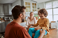 Eine Familie, bestehend aus einer Frau, einem Mann und einem kleinen Jungen, sitzt in der Küche zusammen. Alle lachen. 