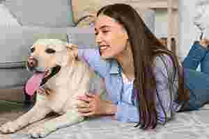Eine junge Frau mit langen braunen Haaren liegt bäuchlings auf dem Fußboden. Neben ihr liegt ein cremefarbener Hund, ein Labrador – sie umarmt den Hund und lächelt ihn liebevoll an.