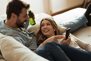 Eine Frau und ein Mann liegen auf einer Couch und kuscheln. Die Frau zeigt dem Mann dabei etwas auf ihrem Handy und der Mann hält eine Flasche in der Hand. Beide teilen sich die Kopfhörer; sie lächeln und wirken sehr zufrieden.