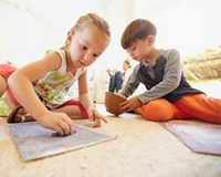 Zwei Kinder malen ein Bild und im Hintergrund sitzt die Mutter.